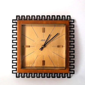 May include: A square wall clock featuring a light wood frame, a gold-toned face with thin black lines for hour markers, and four rectangular markers. The clock has black hands and a black outer border with a repeating geometric Greek key pattern.