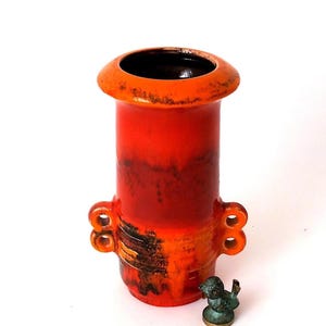 Könnte beinhalten: Eine leuchtend orangefarbene Keramikvase mit einem breiten, ausgestellten Rand und zwei Doppelhenkeln an jeder Seite. Der Körper zeigt einen Farbverlauf aus Rot- und Orangetönen mit dunkelbraunen, strukturierten Akzenten. Eine kleine, verwitterte bronzefarbene Vogelfigur steht daneben.