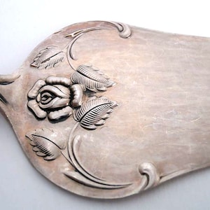 Puede incluir: Servidor de pastel plateado con un diseño floral de rosa y hojas. El servidor tiene una hoja ancha y plana y un mango decorativo. La rosa está en el centro de la hoja, con hojas y vides que la rodean. Probablemente hecho de acero inoxidable.
