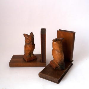 Op de afbeelding: Een paar houten uil-boekensteunen. Elke boekensteun heeft een gebeeldhouwde uil bovenop een stapel boeken. De boekensteunen zijn gemaakt van donkerbruin hout en zijn ontworpen om boeken rechtop op een plank te houden.