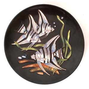 Könnte beinhalten: Ein schwarzer Keramikteller mit einem Reliefdesign von drei Skalaren, die zwischen Seegras schwimmen. Die Fische sind weiß mit schwarzen Streifen und blauen Akzenten. Der Teller hat eine glänzende Oberfläche und ist ein dekorativer Gegenstand.