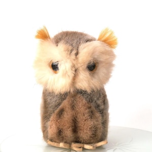 May include: A small, plush owl figurine featuring a soft, fuzzy texture in shades of brown and cream. The owl has large, dark, round eyes, prominent tufted ears with light yellow tips, and a rounded body with a textured, brown chest area.