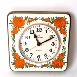 May include: A square Junghans quartz wall clock with a cream-colored face and black numerals. The frame features a decorative border of orange roses with green leaves in each corner. The clock has black hour and minute hands and a thin red second hand.