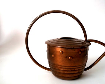 May include: A copper watering can with a rounded body and a handle that forms a circle. The can has a textured surface with small raised dots.