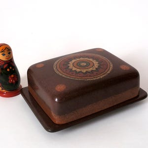 May include: A rectangular brown ceramic butter dish with a decorative circular mandala-style pattern on the lid, accompanied by a small wooden nesting doll featuring a red headscarf and a dark green apron with a painted floral design.
