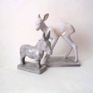 May include: Two ceramic animal figurines, a white deer and a gray donkey, on separate bases. The deer is standing on its hind legs with its head tilted down. The donkey is standing on all fours with its head tilted up. Both figurines are glazed in a white and gray color.