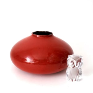 May include: A red, rounded vase with a black interior rim is next to a clear glass owl figurine. The vase has a glossy finish. The owl is detailed with visible eyes and feathers. Both objects are on a white background.