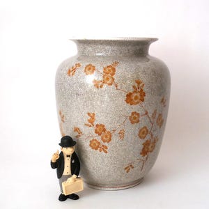 May include: A large, gray ceramic vase with a floral design in shades of orange. The vase has a crackle glaze and a wide, rounded shape. A small figurine of a man in a suit and bowler hat stands in front of the vase.