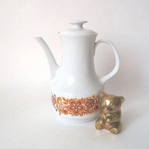 May include: A white ceramic coffee pot with a lid, a curved spout, and a handle. The pot has a decorative floral pattern in shades of orange and brown. A small gold-colored bear figurine sits beside the pot.