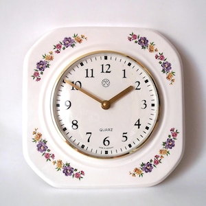 May include: A square white wall clock with rounded corners, featuring a floral pattern of purple and yellow flowers in the corners. The clock face has black Arabic numerals, gold-colored hands, a gold rim, and the text QUARZ and MADE IN GERMANY.