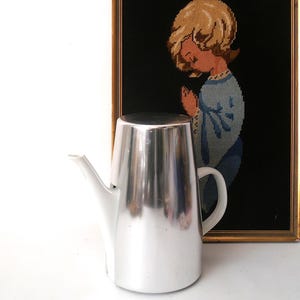 May include: A silver and white coffee pot with a handle and spout. The pot has a shiny, metallic finish. In the background, a framed cross-stitch artwork depicts a child in prayer, with a black background and gold frame.