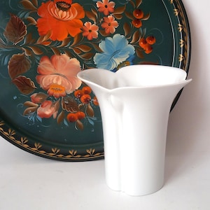 May include: A white ceramic vase with a unique, petal-like top. The vase is set against a dark green tray decorated with a floral design in orange, pink, and blue.