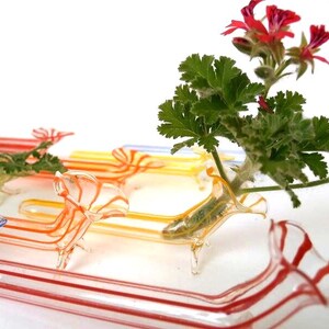 May include: A collection of clear glass bud vases shaped like dachshunds, featuring colorful striped patterns in red, orange, yellow, and blue. One vase holds a green plant with small red flowers, while others are arranged in the foreground and background.