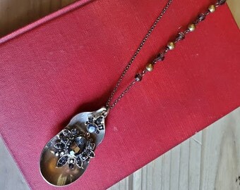 May include: A silver spoon necklace with black and clear rhinestones, hanging from a brown chain on a red book.