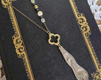 May include: A vintage necklace featuring a gold-coloured clover pendant and a silver spoon-shaped charm. The necklace has a delicate chain and is decorated with cream and iridescent beads. The background is a dark book with gold decorative edges.