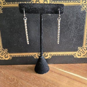 May include: A pair of silver-colored, twisted-design earrings hanging from a black jewelry display stand. The earrings are long and slender, with a textured, rope-like appearance. The background is a dark, ornate book cover.