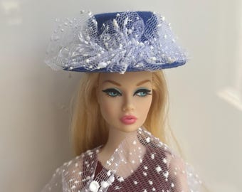 Könnte beinhalten: Ein blauer Puppenhut mit weißer Spitze und Tupfen. Der Hut befindet sich auf einer Puppe mit blonden Haaren, blauen Augen und rosa Lippen. Die Puppe trägt ein weinrotes Kleid und einen weißen Schal mit Tupfen.