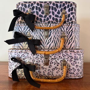 May include: A stack of three rectangular storage cases with rounded corners, featuring animal print patterns in shades of pink, purple, and black. Each case has a bamboo-style handle, a silver metal latch, and a black ribbon tied to the handle.
