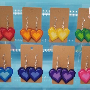 May include: Eight pairs of pixel heart earrings displayed on brown card stock. The earrings are in the colors red, orange, yellow, green, blue, purple, and pink.
