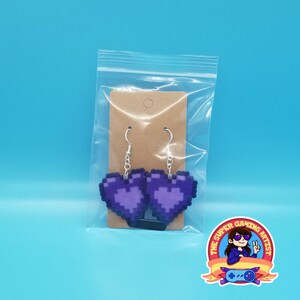 May include: A pair of purple pixel heart earrings on a brown card with a hole for hanging. The earrings are packaged in a clear plastic bag.