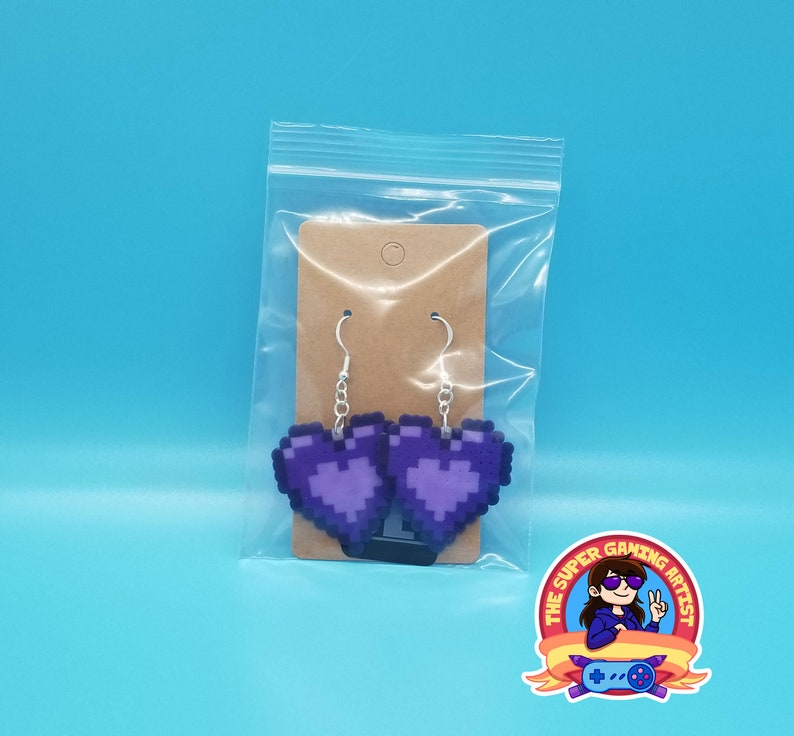 May include: A pair of purple pixel heart earrings on a brown card with a hole for hanging. The earrings are packaged in a clear plastic bag.