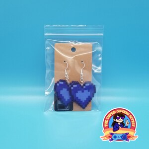 May include: A pair of purple pixel heart earrings with silver hooks. The earrings are packaged in a clear plastic bag with a brown card backing.