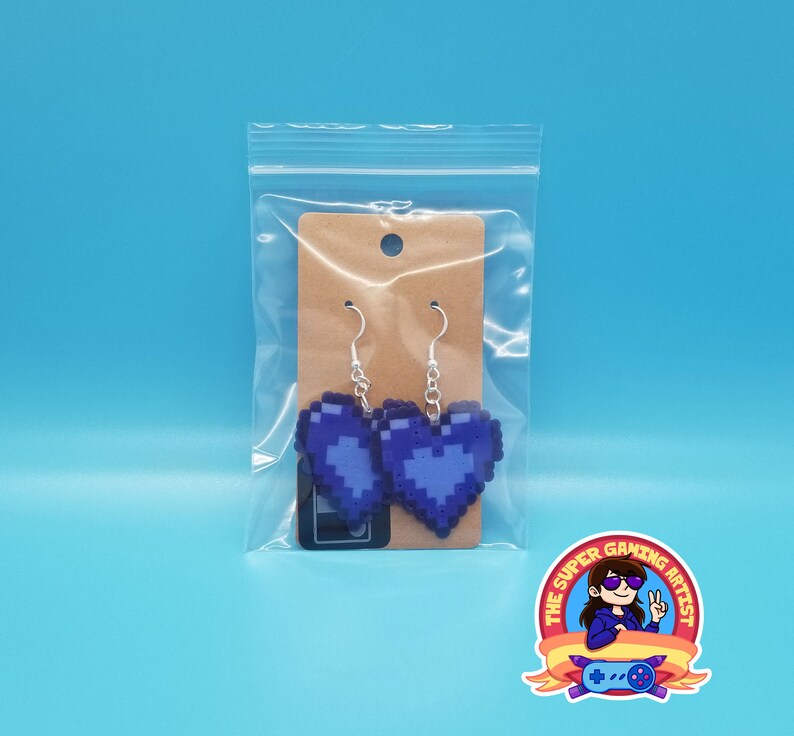 May include: A pair of purple pixel heart earrings with silver hooks. The earrings are packaged in a clear plastic bag with a brown card backing.