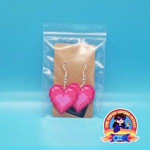 May include: Pink pixel heart shaped earrings in a clear plastic bag with a brown paper tag. The earrings are on a white background. The Super Gaming Artist logo is in the bottom right corner.