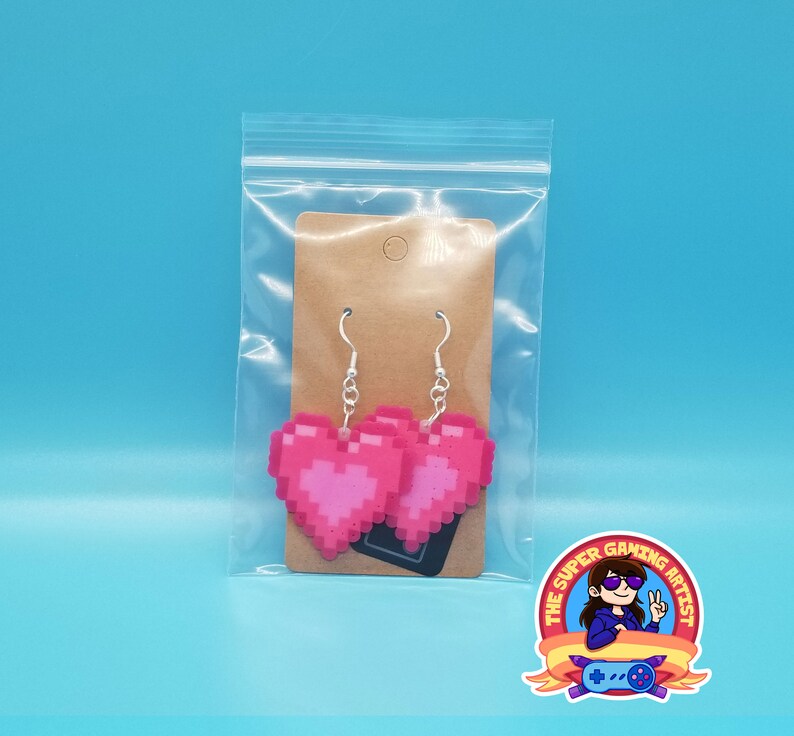 May include: Pink pixel heart shaped earrings in a clear plastic bag with a brown paper tag. The earrings are on a white background. The Super Gaming Artist logo is in the bottom right corner.