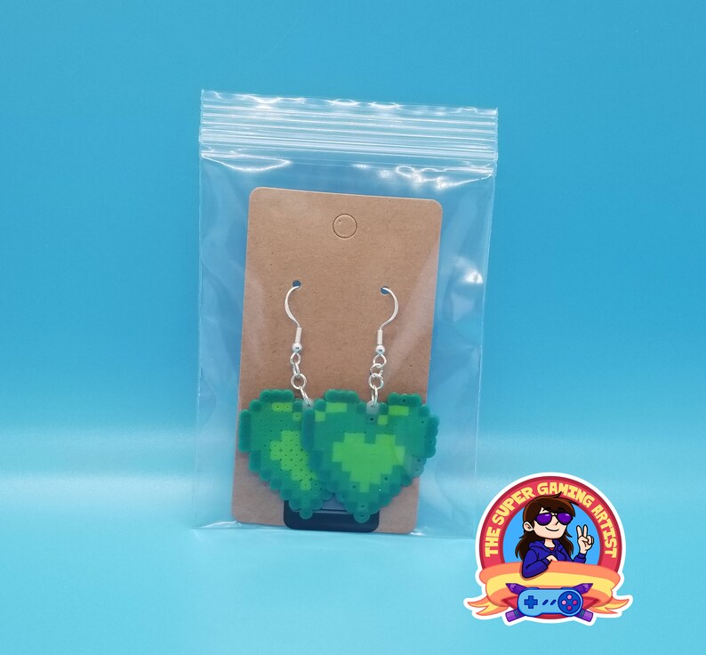 May include: A pair of green pixel heart earrings on a brown card with a white hole in the top. The earrings are hanging from silver hooks. The card is in a clear plastic bag. The logo "The Super Gaming Artist" is visible in the bottom right corner of the image.