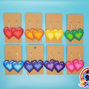 May include: Eight pairs of pixel heart earrings on brown card stock. The hearts are in rainbow colors: red, orange, yellow, green, blue, purple, and pink.