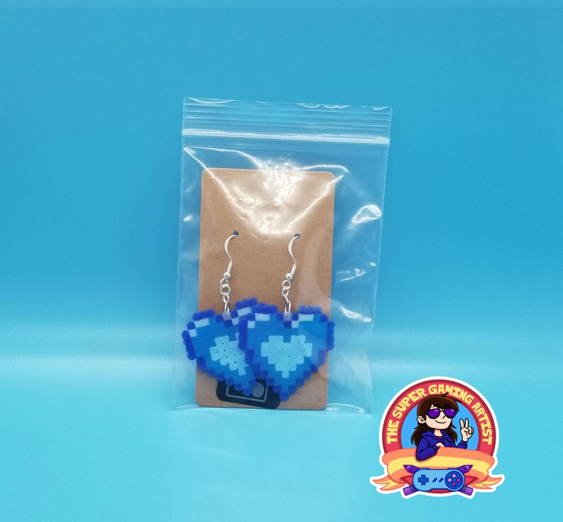 May include: A pair of blue pixel heart earrings in a clear plastic bag with a brown card backing. The earrings are on silver hooks. The bag has a sticker on the front that says "The Super Gaming Artist".