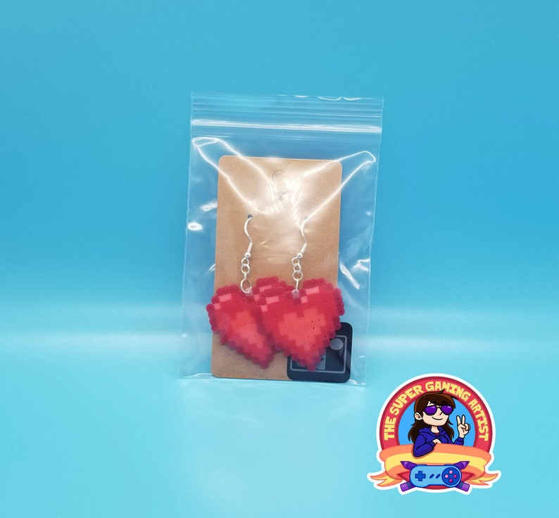 May include: A pair of red pixel heart earrings on a brown card with a white background. The earrings are hanging from silver hooks.