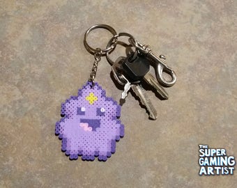 May include: A purple pixel art keychain featuring a cartoon character with a yellow star on its head, attached to a keyring with keys and a clip. The keychain is made of small, colorful beads. The Super Gaming Artist logo is in the bottom right corner.