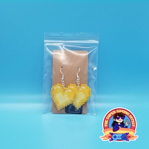 May include: A pair of yellow pixel heart earrings on a brown card in a clear plastic bag. The earrings are on silver hooks.