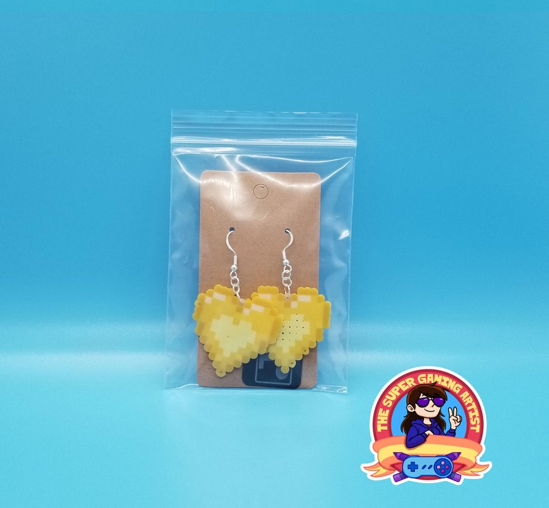 May include: A pair of yellow pixel heart earrings on a brown card in a clear plastic bag. The earrings are on silver hooks.