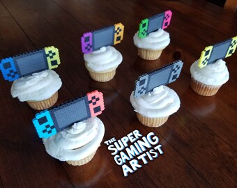 May include: Six cupcakes decorated with colorful pixel art Nintendo Switch controllers. The cupcakes are on a wooden table. The text "The Super Gaming Artist" is visible in the bottom right corner of the image.