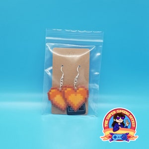 May include: A pair of orange pixel heart earrings on a brown card in a clear plastic bag. The earrings are on silver hooks. The card has a small black sticker with the text "The Super Gaming Artist".
