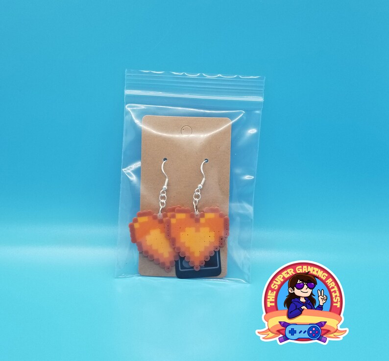 May include: A pair of orange pixel heart earrings on a brown card in a clear plastic bag. The earrings are on silver hooks. The card has a small black sticker with the text "The Super Gaming Artist".