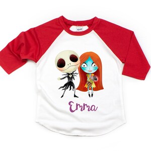 Puede incluir: Camiseta de b&eacute;isbol blanca y roja con una imagen de dibujos animados de Jack Skellington y Sally de Pesadilla antes de Navidad. La camiseta tiene el nombre "Emma" impreso en morado debajo de la imagen.
