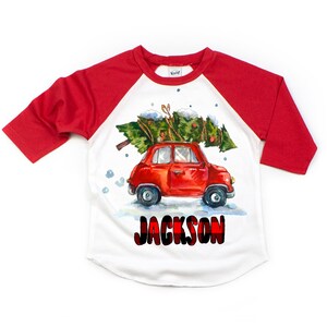 May include: A white and red baseball tee shirt with a red car carrying a Christmas tree. The text "JACKSON" is printed in black with a red and black plaid pattern.