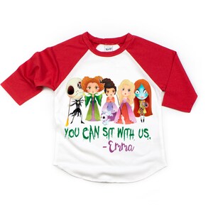 Puede incluir: Una camiseta de b&eacute;isbol blanca y roja con un gr&aacute;fico de personajes de dibujos animados de pel&iacute;culas populares de Halloween. El texto "You can sit with us. -Emma" est&aacute; impreso en la camiseta.