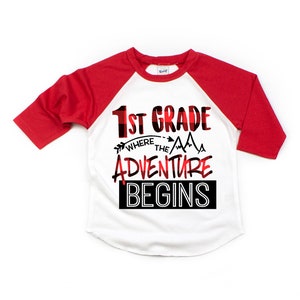 May include: A white and red baseball tee shirt with a black and red plaid design. The shirt has the text "1st Grade Where the Adventure Begins" printed on the front.