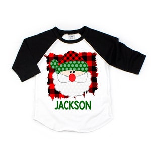 May include: A white and black baseball tee shirt with a red and green plaid Santa Claus face graphic. The text "JACKSON" is printed in green below the graphic.