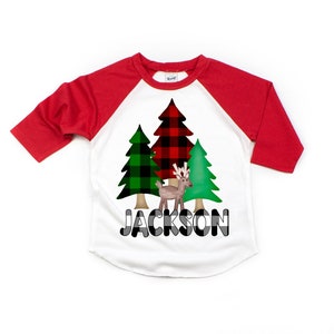 May include: A white and red baseball tee shirt with a green and black plaid Christmas tree design and a reindeer. The shirt says "JACKSON" in black and white plaid letters.