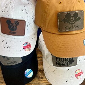 May include: Four baseball caps with black and white splatter paint designs. Each cap has a personalized leather patch with a Mickey Mouse design and a name. The names on the patches are Jordan, Asher, One Cool Ghoul, and Asher.