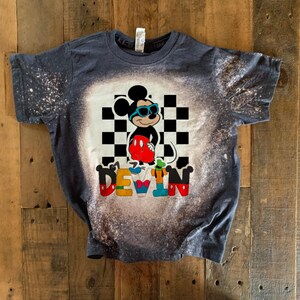 May include: A dark blue t-shirt with a bleach-splattered design featuring Mickey Mouse wearing sunglasses and a checkered background. The name "DEVIN" is spelled out below Mickey Mouse in colorful letters.