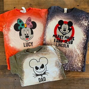 Puede incluir: Tres camisetas blanqueadas con diferentes dise&ntilde;os de tem&aacute;tica de Halloween. La primera camiseta es naranja con un dise&ntilde;o de Mickey Mouse en blanco y negro y el texto "LUCY". La segunda camiseta es azul marino con un c&iacute;rculo rojo alrededor de un dise&ntilde;o de Mickey Mouse en blanco y negro y el texto "MICKEY MOUSE FANG CLUB LINCOLN". La tercera camiseta es verde con un dise&ntilde;o de Jack Skellington en blanco y negro y el texto "DAD".