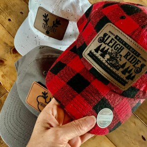 May include: Three baseball caps with different designs. One is white with black speckles and a brown leather patch that says "Theodore". Another is red and black plaid with a brown leather patch that says "Main Street Sleigh Rides". The third is gray with a brown leather patch that says "Theodore".