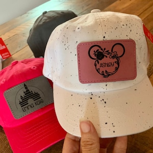 Pu&ograve; includere: Due cappellini da baseball personalizzati. Uno &egrave; rosa con una grafica di un castello bianco e il testo "Emma Kate". L'altro &egrave; bianco con macchie nere e una toppa rosa con una silhouette di Topolino nera e il testo "Ashley".